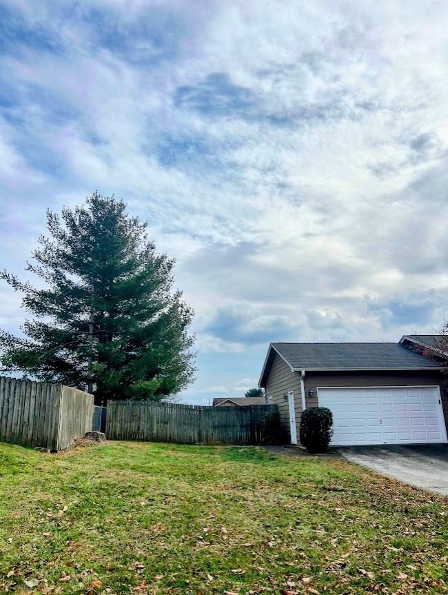 A side view of a house with a garage and a green lawn, featuring a tall pine tree and a wooden fence under a cloudy sky.